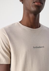 Tricou bej cu mânecă scurtă, fabricat dintr-un material moale și ușor, având o mică imprimare „Arrivederci” de culoare gri în zona stângă a pieptului.