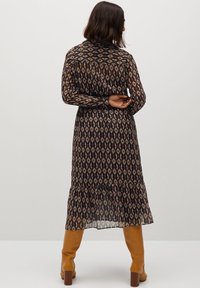 Vestido largo y transparente de estampado oscuro con patrones geométricos. Con dobladillo con volantes, acentos en marrón oscuro y rojo. Se lleva con botas de caña alta color beige.