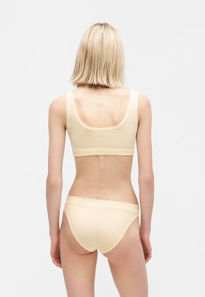 Γυναίκα με ξανθά μαλλιά που φοράει μπεζ αθλητικό σουτιέν και αντίστοιχο εσώρουχο, φαίνεται από πίσω με φόντο ένα απλό γκρι φόντο.