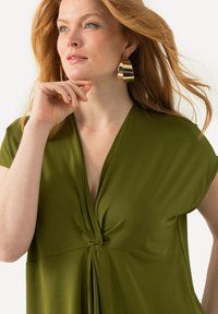 Olivově zelený top s hlubokým výstřihem do V a zkrouceným středovým detailem. Krátké rukávy a hladká textura látky zvýrazňují design. Zlaté kruhové náušnice.