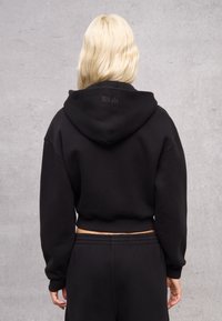 Sudadera negra recortada hecha de tela suave, con capucha ajustable y logo en relieve en la parte posterior. Corte holgado con mangas largas.