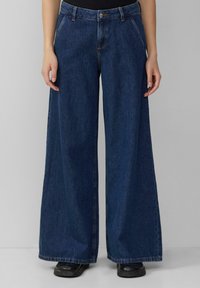Jean en denim bleu foncé à jambes larges avec taille haute, fermeture à bouton à l'avant et détails de couture visibles.