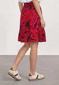 Femme portant une jupe fleurie rouge et des baskets blanches avec des accents marron, debout sur un sol gris contre un mur blanc.