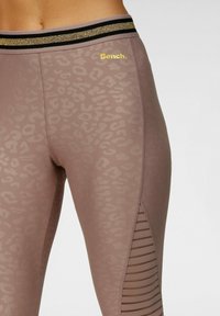 Leggings in taupe, con un sottile motivo leopardato e una fascia in vita a righe nere e dorate. Pannello laterale con aperture verticali per migliorare la ventilazione.