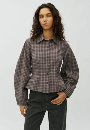 Young woman with curly hair wears a fitted, long-sleeve, brown and gray checkered button-up shirt and dark pants against a plain background.