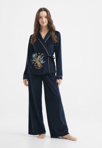 Conjunto de pijama de terciopelo azul marino con blusa de manga larga tipo bata que presenta ribetes en rosa y bordados florales. Los pantalones de pierna ancha completan el look.