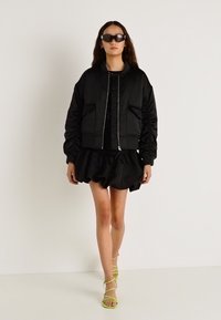 Fekete bomberdzseki bordázott mandzsettákkal, fekete fodros szoknyával és sárga pántos magas sarkúval párosítva. A modell sötét napszemüveget visel.