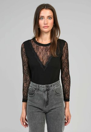 Femme portant un haut en dentelle noire à manches longues avec un motif floral et un jean gris taille haute, se tenant devant un fond gris uni.