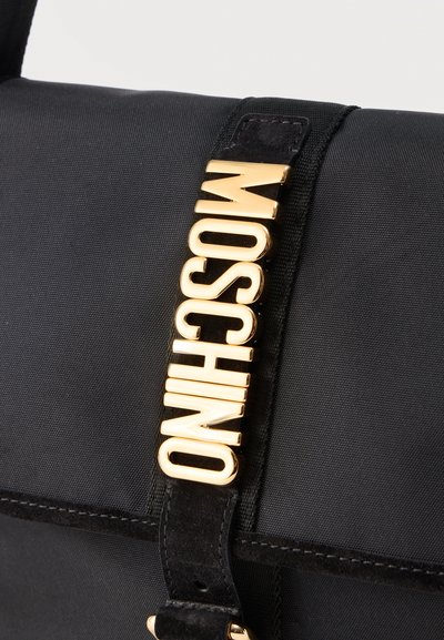 Μαύρη υφασμάτινη τσάντα χειρός με μαύρο λουρί που φέρει χρυσά γράμματα που σχηματίζουν κάθετα τη λέξη "MOSCHINO".