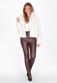 Kvinde iført en hvid faux fur jakke, cremefarvet top, maroon læderbukser og sorte spidse heels, der smiler mod en hvid baggrund.