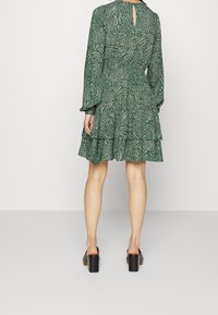 Vihreä kuvioitu mekko pitkillä hihoilla, kerroksittain rypytetty helma ja takana avainnauhat yksityiskohtana, yhdistettynä mustiin palakorkoisiin kenkiin.