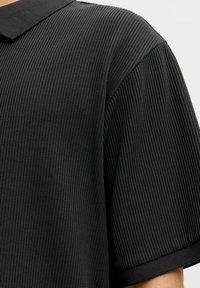 Gros plan sur un t-shirt à manches courtes côtelé foncé montrant le tissu texturé et la couture d'épaule sur une personne devant un fond clair.