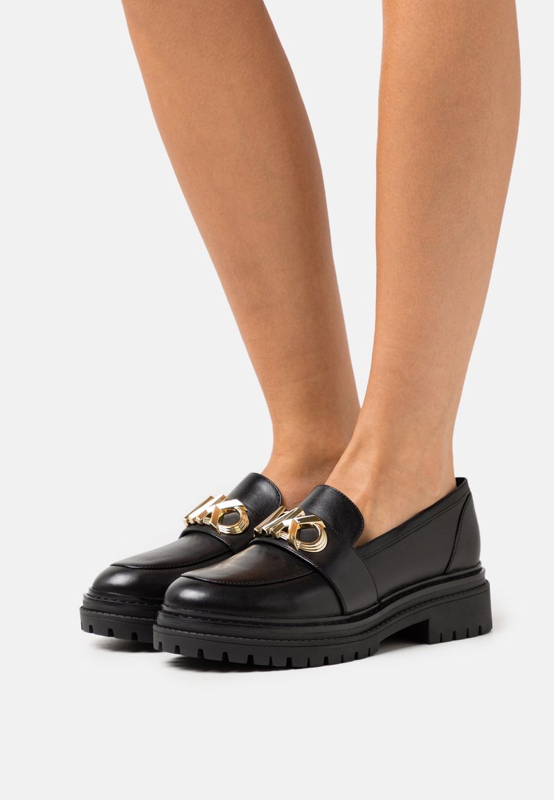 Mocassins en cuir noir avec semelles épaisses à crampons et grand détail de chaîne dorée portés sur pieds nus sur un fond uni.