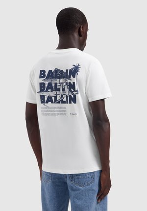 Man draagt een wit T-shirt met een marineblauwe afbeelding van een huis en een palmboom en herhaalde tekst "BALLIN" op de achterkant, gecombineerd met een blauwe spijkerbroek.