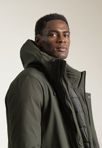 Chaqueta aislante de color verde oscuro con un alto cuello y capucha desmontable, que presenta un cierre con cremallera y bolsillos exteriores. Acabado suave y texturizado.