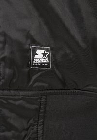 Črn material z gladko teksturo, na katerem je prišit črn etiketa z belim logotipom "STARTER" in besedilom "BLACK LABEL".