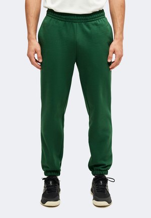 Homme portant un pantalon de jogging vert foncé à taille élastique avec des baskets noires, debout, les bras détendus le long du corps, sur un fond uni.