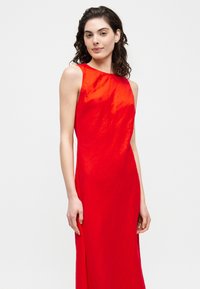 Γυναίκα με σγουρά σκούρα μαλλιά, ντυμένη με αμάνικο κόκκινο φόρεμα, στέκεται μπροστά σε απλό ανοιχτό φόντο.