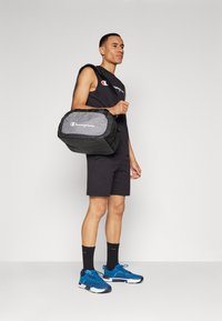 Bolsa de sport negra y gris con el logo de "Champion", tela suave, asa superior para llevar, y correa de hombro ajustable. Modelo llevando una camiseta sin mangas.