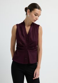 Blouse sans manches en bordeaux profond avec un col en V, des détails de taille froncée et une fermeture à bouton. Tissu doux avec une coupe ajustée.