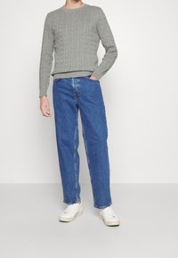 Šedý svetr s klasickým vzorem copánkového pletení a zaobleným výstřihem, doplněný volnými modrými džínami a bílými teniskami. Jednoduchý a ležérní design.