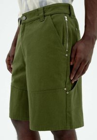 Olive green shorts made of sturdy fabric, featuring a side zipper pocket and snap-button accents, with a straight cut and knee-length design.