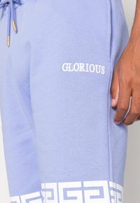 Světlé fialové tepláky vyrobené z měkké tkaniny, s bílým vyšitým nápisem "GLORIOUS" a geometrickým bílým vzorem na lemu.