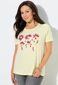 Żółty t-shirt z krótkim rękawem, zdobiony fioletowymi stokrotkami i żółtymi sercami w kwiatowy wzór. Miękka tkanina z okrągłym dekoltem. Szare dżinsy.