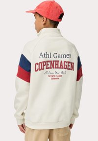 Sudadera de marca Copenhague en color crema, con mangas a rayas rojas y azules, un cuello alto y texto en negrita en la parte posterior. Textura de tela suave.