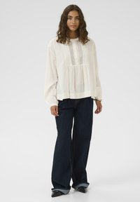 Femeie cu păr ondulat castaniu, purtând o bluză albă lungă cu mâneci largi, blugi negri largi și pantofi negri cu vârful ascuțit, stând în fața unui fundal simplu.