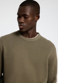 Texturēts olīvzaļš adīts džemperis ar ribinātu apaļu izgriezumu. Tuvplāns uzsver auduma sarežģīto rakstu un slāņošanu.