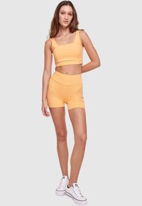 Orange crop top med fyrkantig halsringning, matchande högmidjade shorts. Modellen bär vita sneakers och står på en neutral bakgrund.