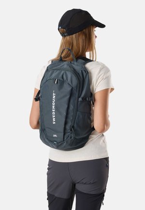 Kobieta w czapce z daszkiem, beżowej koszulce i szarych spodniach z ciemnoszarym plecakiem Swedemount 20L na plecach, zwrócona tyłem, na białym tle.
