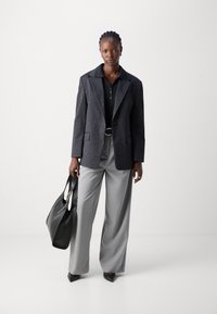 Blazer noir à rayures sur une polo noire, pantalon gris à jambes larges, associé à un sac tote noir et des talons noirs pointus.