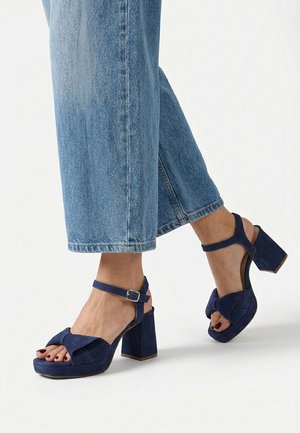Pies con sandalias de tacón cuadrado de ante azul con tiras en el tobillo, combinadas con vaqueros azules recortados.