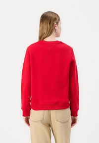 Rød genser med ribbestrikkede mansjetter og nederkant, med rund hals og synlige sømdetaljer. Kombinert med lysebeige bukser.