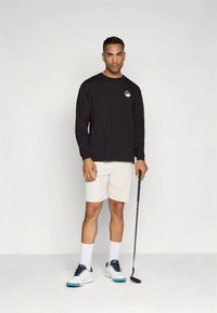 Ilgas rankovėmis juodas marškinėlis su grafiniu logotipu, baltos šortai, balti sportbačiai su mėlynais akcentais ir golfo lazda rankoje.