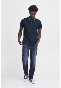Marineblå polo t-shirt med kontrastfarvet krave, parret med slidte blå jeans og sorte sneakers. Den har et logo på brystet og korte ærmer.