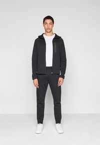 Felpa nera con cappuccio e zip frontale con tasca davanti, abbinata a pantaloni affusolati dello stesso colore e sneakers bianche. T-shirt bianca semplice sotto.