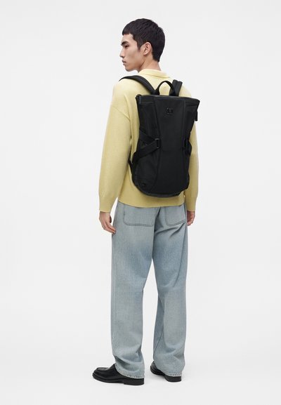Personne portant un pull jaune clair, un jean bleu clair, des chaussures noires et un grand sac à dos noir, debout devant un fond blanc uni.