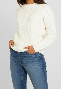 Frau in einem weißen Kabelstrickpullover mit strukturierten Mustern und blauen, eng anliegenden Jeans, die Hände in den vorderen Taschen.