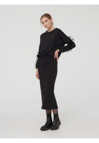 Felpa nera con dettagli in volant trasparenti sulle maniche, abbinata a una gonna nera aderente, indossata con stivali neri spessi alla caviglia.