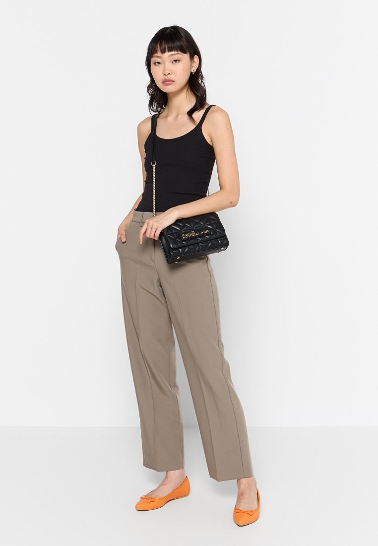 Mujer vistiendo una camiseta negra de tirantes, pantalones beiges, zapatos planos naranjas y sosteniendo un bolso cruzado negro acolchado con cadena dorada.
