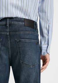 Mørkeblå denimjeans med en glat tekstur, der har et waistband-label i sort læder, baglommer og en tætsiddende silhuet.
