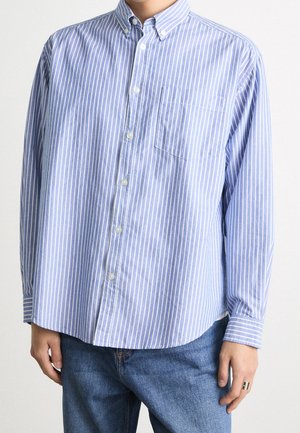 Personne portant une chemise à rayures verticales bleues et blanches avec manches longues et poche poitrine, associée à un jean bleu.