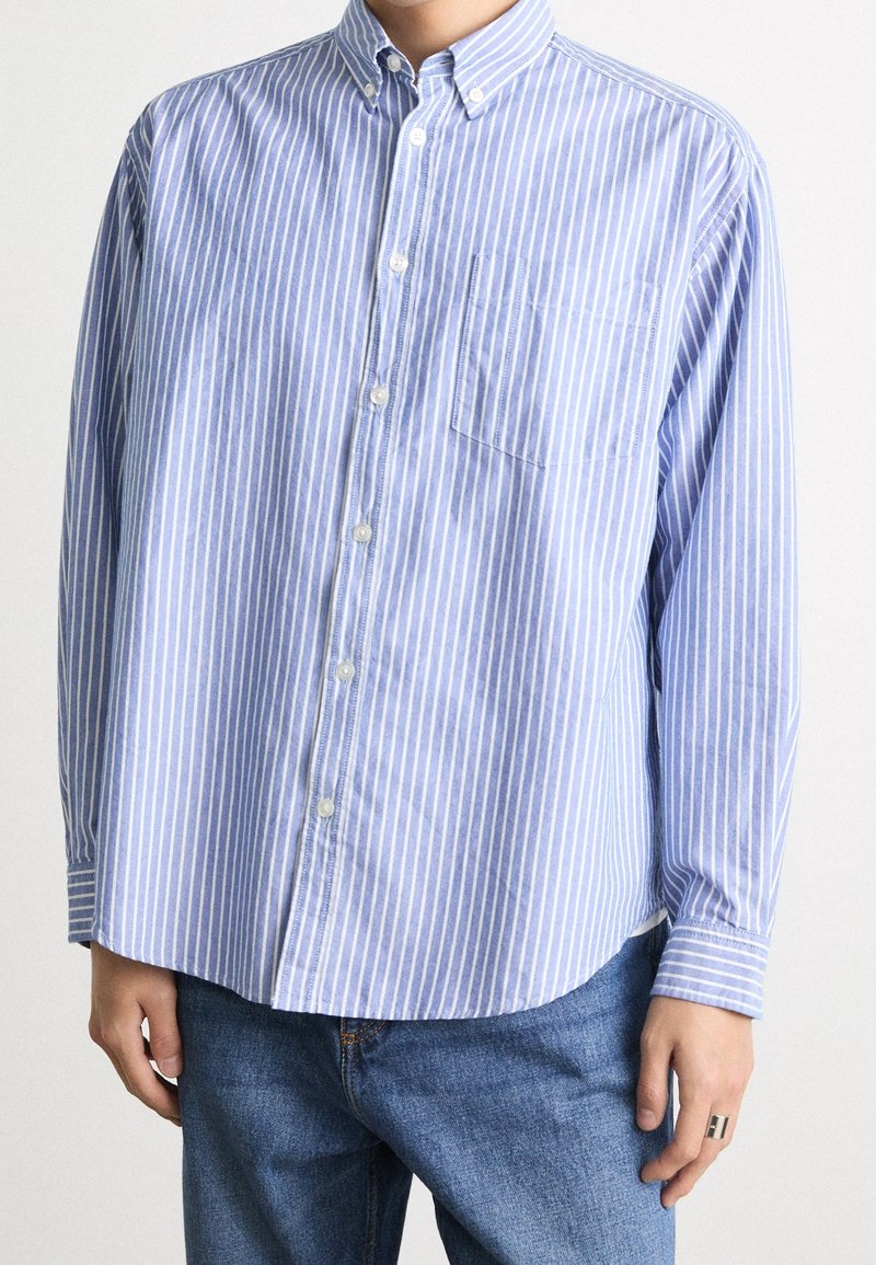 Personne portant une chemise à rayures verticales bleues et blanches avec manches longues et poche poitrine, associée à un jean bleu.