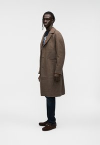 Καφέ μάλλινο παλτό με κλασικό σχεδιασμό, γιακά με πέτο, δύο μπροστινές τσέπες· συνδυασμένο με σκούρο τζιν και καφέ σουέντ παπούτσια.