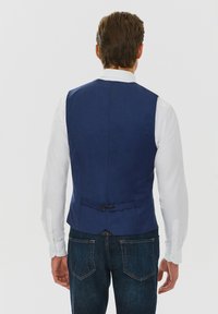 Granatowa kamizelka z teksturowanej tkaniny, z tylnym ściągaczem i dwoma czarnymi guzikami, noszona na białej bluzce z długim rękawem i ciemnych dżinsach.