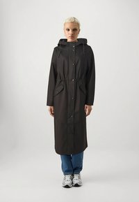 Długi czarny płaszcz przeciwdeszczowy z kapturem, ściągaczem w talii i zapięciem na zatrzaski, wyposażony w kieszenie oraz gładkie, matowe wykończenie. W połączeniu z niebieskimi dżinsami i trampkami.