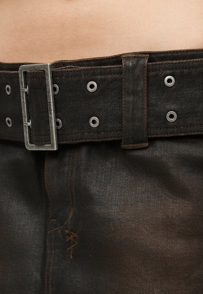 Gros plan sur un pantalon en denim noir avec une large ceinture noire dotée d'une boucle métallique rectangulaire et d'œillets en métal, portée à la taille.